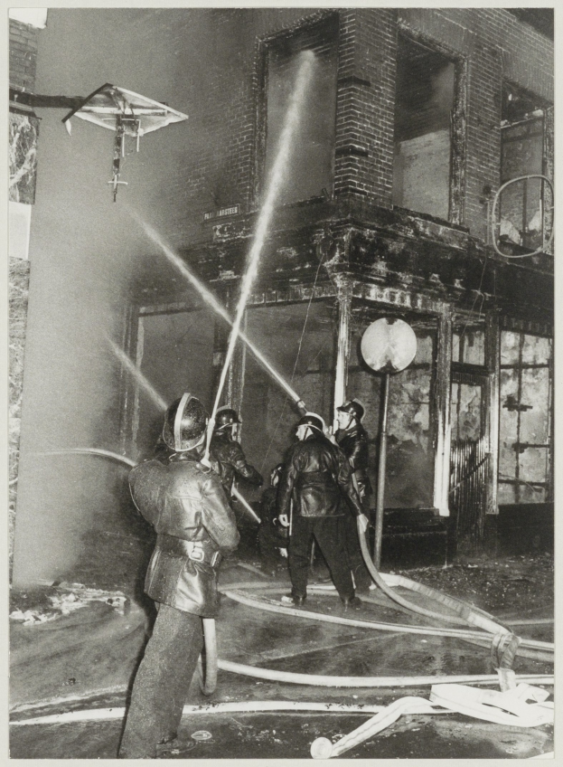 Schwarzes und weißes Foto von Feuerwehrmännern in Helmen, die Schläuche vor einem brennenden Gebäude halten.