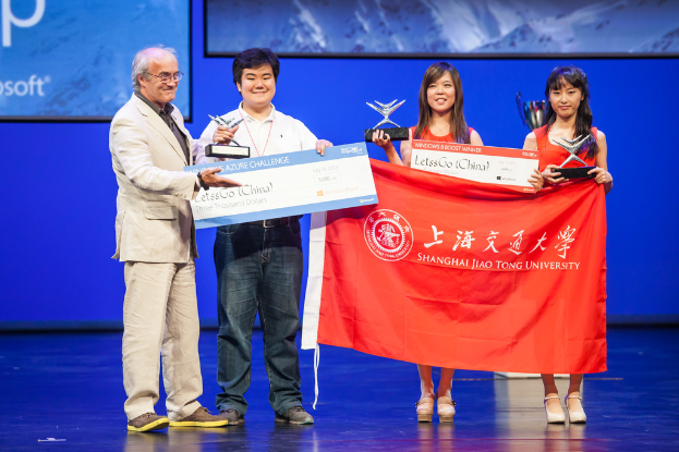 Vier Personen stehen auf einer Bühne, jede hält Auszeichnungen, Schecks und eine Fahne, sie lächeln, mit einem digitalen Bildschirm im Hintergrund.