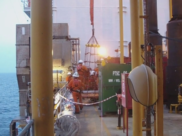 Ein Schiff mit Masten, Kisten und Geländern, mit Menschen in Helmen, vor Wasser.