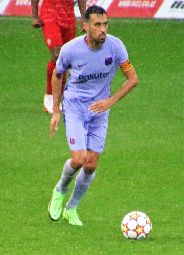 Ein Fußballspieler in einem Sportoutfit und -schuhen ist im Einsatz auf dem Feld, wobei ein Textschild im Hintergrund sichtbar ist.