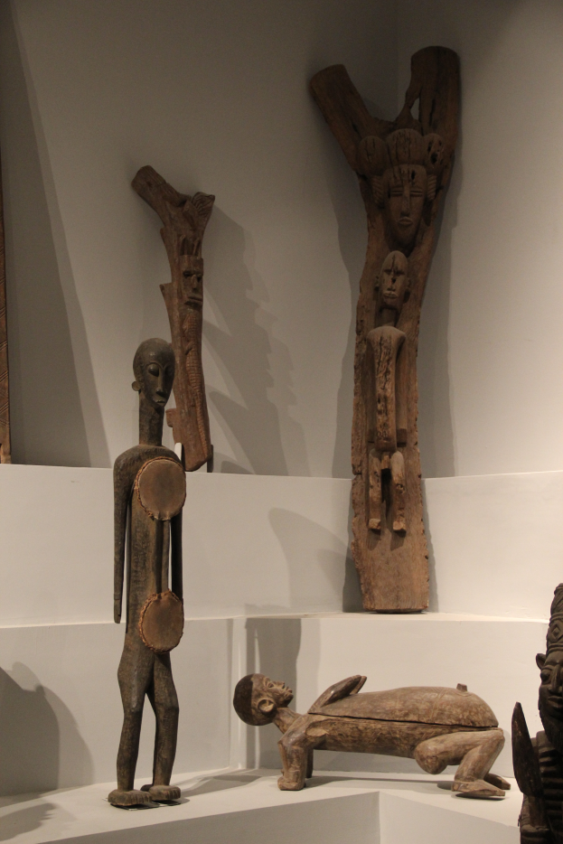 Eine Gruppe von Holzskulpturen, die auf einer weißen Fläche in einem Museum ausgestellt sind, mit einer Wand im Hintergrund.