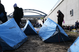 Eine Gruppe von Menschen sitzt auf einem sandigen Strand neben Zelten, die besorgt aussehen, mit einer Wand auf der rechten Seite und einer Brücke im Hintergrund.