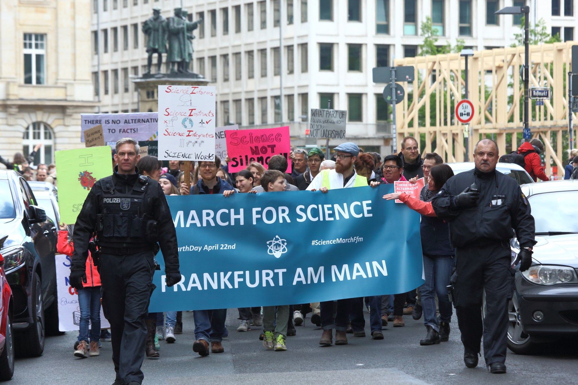 Gruppe von Menschen marschiert auf der Straße mit einer "March for Science Frankfurt am Main"-Fahne vorbei, Autos und Gebäude im Hintergrund.