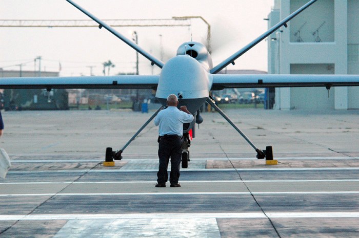 RQ-1 Predator Unmanned Aerial Vehicle auf einem Flughafenvorfeld mit zwei Personen in der Nähe, vor Buildings, Bäumen, Polen und einem klaren blauen Himmel.