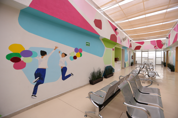 Ein Krankenhausflur mit Stühlen auf der rechten Seite, ein buntes Wandgemälde, Topfpflanzen, eine Tür, ein Schild und eine Decke mit Lampen.