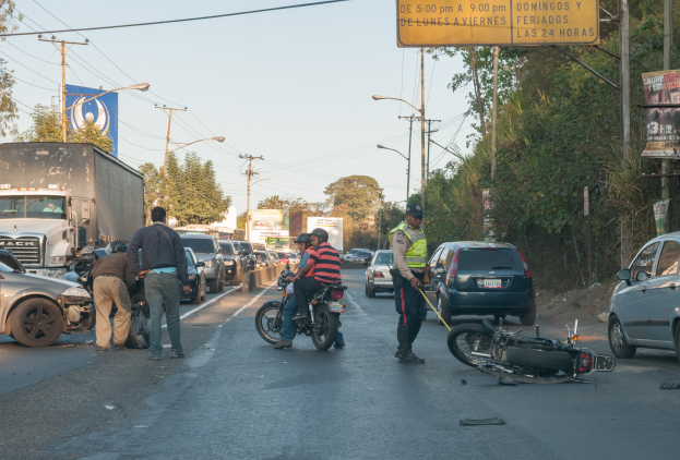 Eine Gruppe von Menschen steht um ein verunglücktes Motorrad auf der Straße herum, umgeben von mehreren Fahrzeugen, einschließlich eines Lastwagens, und einer Hintergrundlandschaft mit Bäumen, Pfosten, Lampen und Schildern unter dem Himmel.