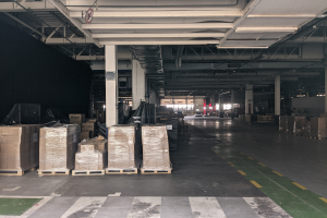 Großer Lagerraum mit zahlreichen Kartonagen auf Paletten, gestützt von Säulen und beleuchtet von Deckenlampen, mit einem an der Decke angebrachten Schild, das ein Flottenmanagementsystem anzeigt.