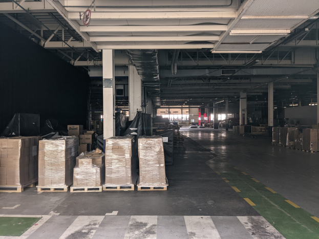 Großer Lagerraum mit zahlreichen Kartonagen auf Paletten, gestützt von Säulen und beleuchtet von Deckenlampen, mit einem an der Decke angebrachten Schild, das ein Flottenmanagementsystem anzeigt.