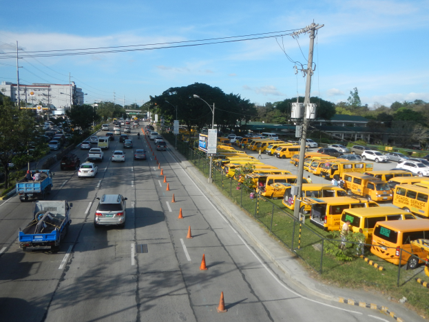 Eine belebte Straßenszene mit verschiedenen Fahrzeugen, Verkehrskegeln, Laternenmasten, Strommasten mit Drähten und Texttafeln, mit Maschendrahtzaun auf der rechten Seite, Bäumen, Gebäuden und einem bewölkten Himmel im Hintergrund.