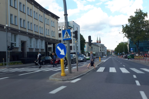 Eine Stadtstraßenszene mit Fußgängern an einer Ampel, Fahrzeugen auf der Straße, Verkehrszeichen, Schildern, Laternen, Bäumen, Gebäuden mit Fenstern und einem bewölkten Himmel.