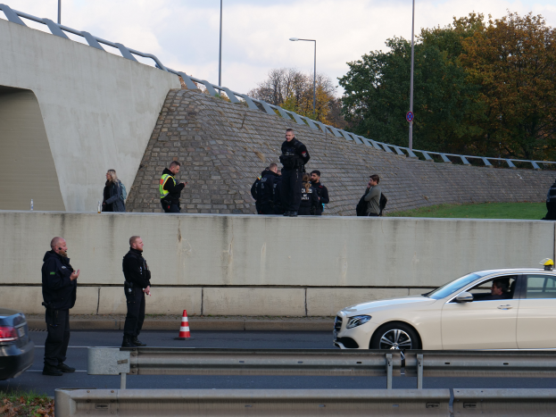 Eine Gruppe von Polizisten steht neben einem Auto auf der Straßenseite, mit Verkehrskegeln, einer Trennwand, Gras, einer Wand, Laternen, Bäumen und einem bewölktem Himmel im Hintergrund.