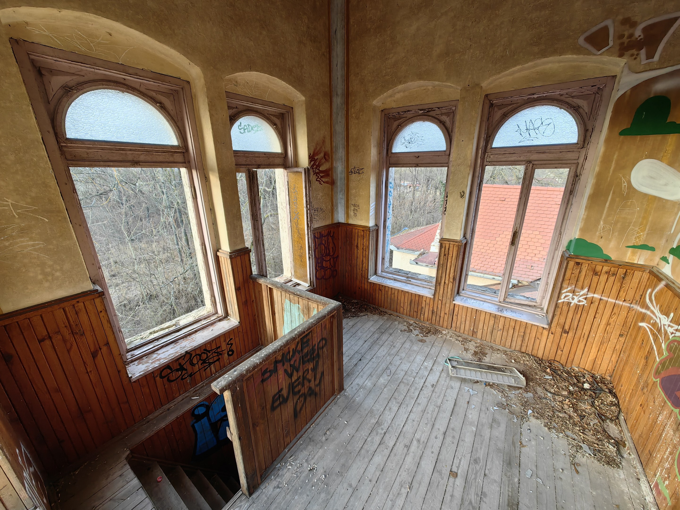 Verlassenes Gebäude mit Graffiti an den Wänden und Fenstern, einem Holzfußboden, einer Treppe und verstreuten Gegenständen, vor einem Hintergrund aus Bäumen und einem fernen Haus.