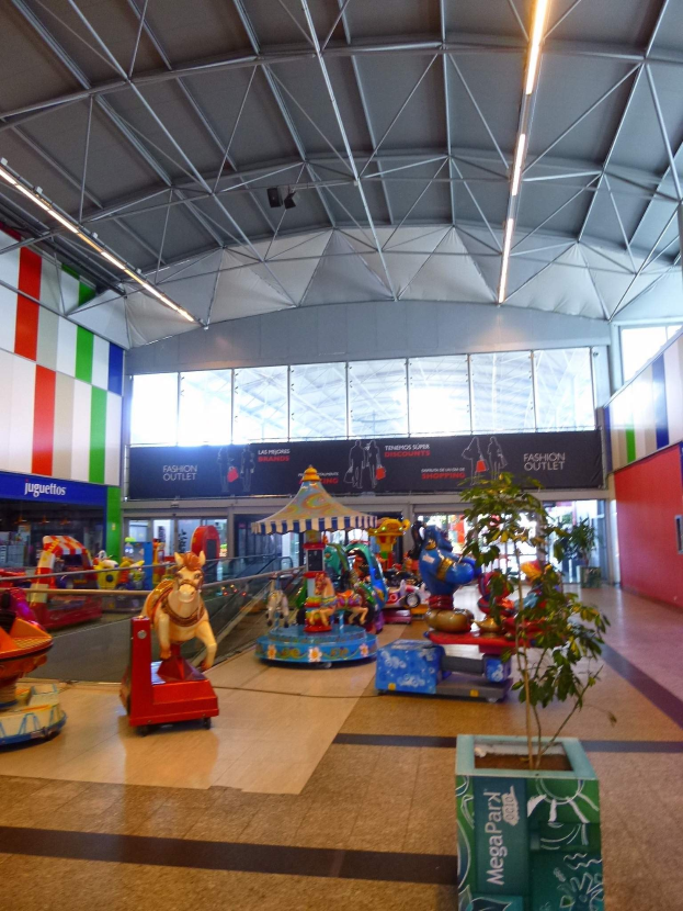 Großer Indoor-Vergnügungspark in einem Einkaufszentrum mit verschiedenen Fahrgeschäften, einem Topf mit einer Pflanze, verstreuten Spielzeugen, Informationsschildern, Deckenleuchten und Metallstützpfosten.