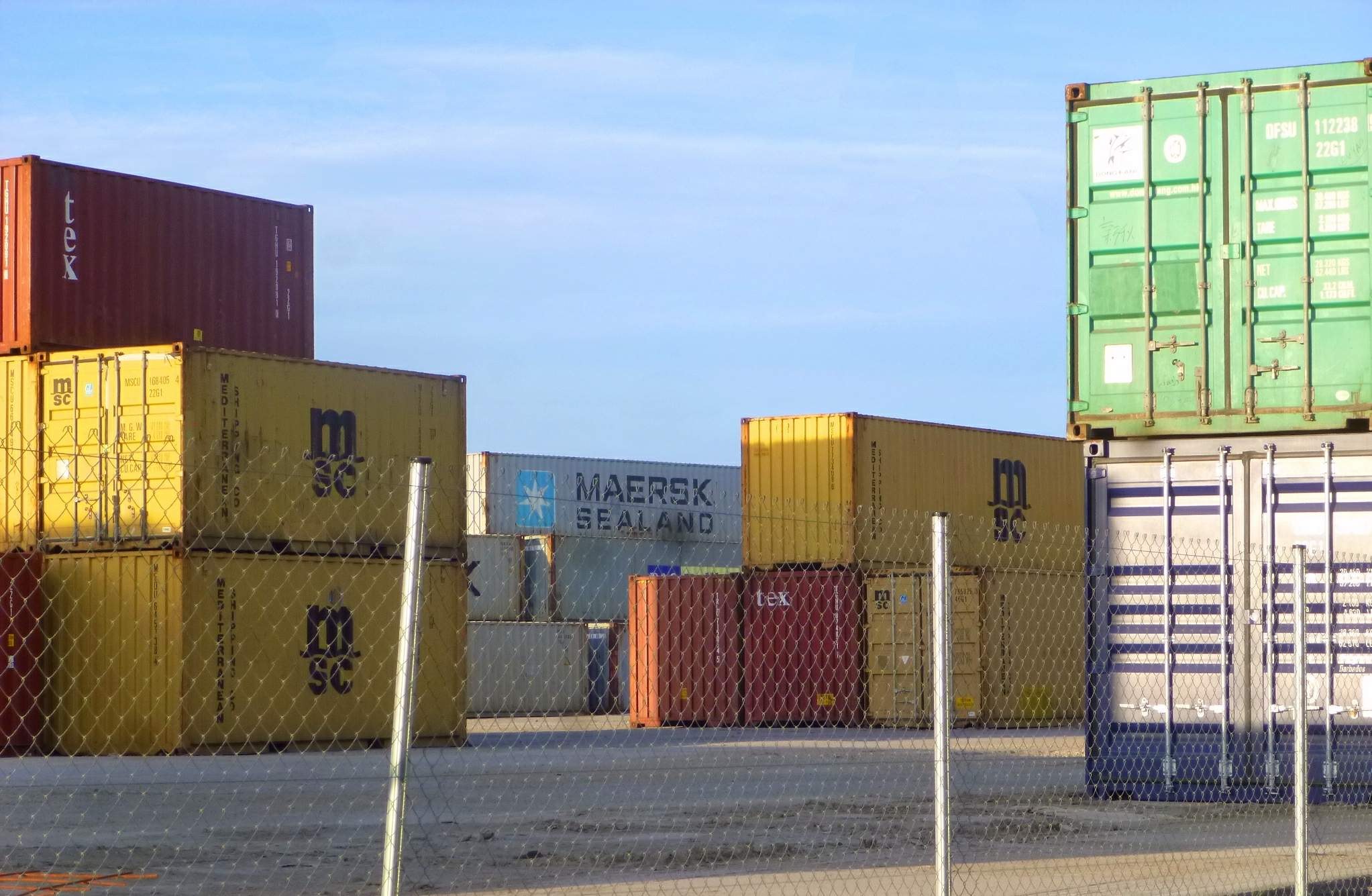Ein hoher Stapel von Containern mit Maschendrahtzaun im Vordergrund, eine Straße unten und ein bewölkter Himmel im Hintergrund.