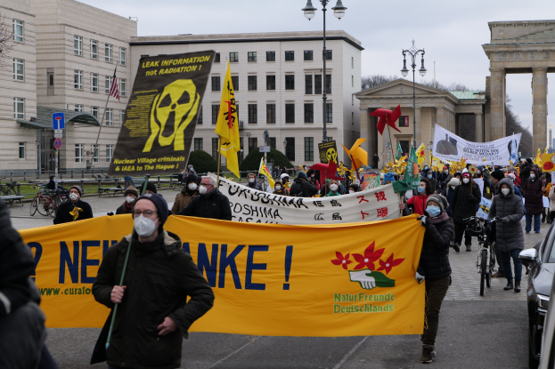 Große Gruppe von Menschen bei einer Straßen-demo gegen Atomkraft in Deutschland, mit Schildern und Fahnen, sowie Fahrzeugen und Gebäuden im Hintergrund.
