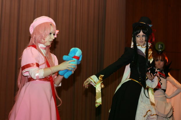 Drei Frauen führen ein Drama auf, wobei eine in einem rosafarbenen Kleid mit einem blauen Teddybär und eine andere in einem schwarzen Kleid, die nach links schaut, zu sehen ist.