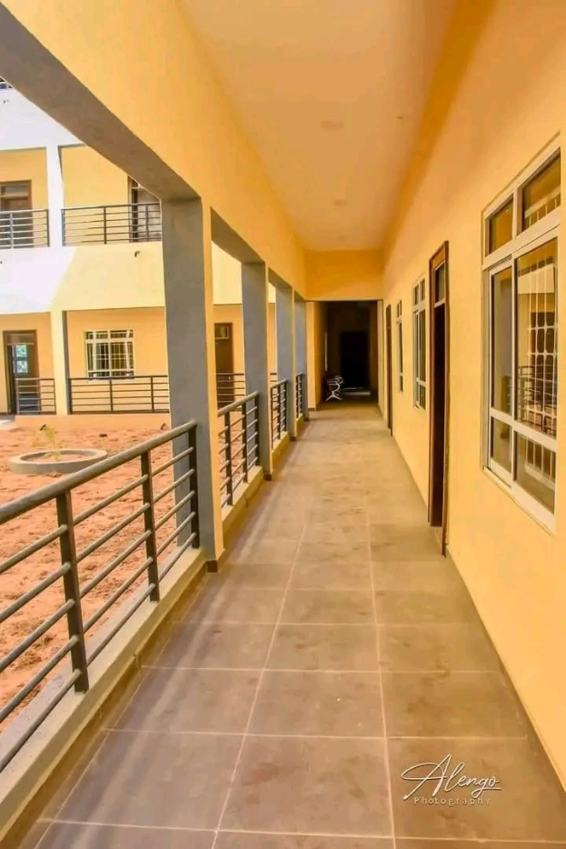 Ein mehrstöckiges Gebäude mit Fenstern, Türen, Säulen und Geländern, das einen langen gefliesten Flur, einen Balkon und andere Gebäude unter einem klaren blauen Himmel zeigt, mit Text, der eine 3-Zimmer-Wohnung in Ntinda, Kampala, zur Miete anbietet.