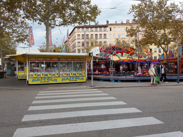 Eine belebte Stadtstraße mit Zebrastreifen und Karussell, mit Menschen, Buden, Bäumen, Gebäuden, Bannern, Laternenmasten und einer Ampel unter einem bewölkten Himmel.