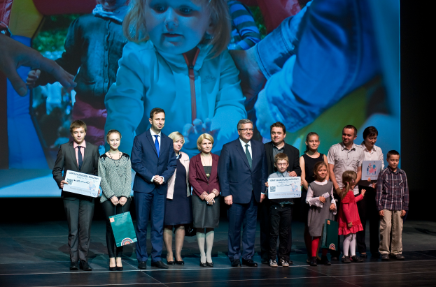 Eine Gruppe von Menschen auf einer Bühne, einige halten Taschen und Urkunden, mit einem Bildschirm im Hintergrund, der ein Kind und andere Personen anzeigt.