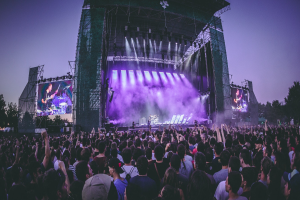 Eine große Menge bei einem Musikfestival schaut auf der Bühne mit Instrumenten und Bildschirmen, die vor einem Gebäude, Bäumen und einem blauen Himmel beleuchtet sind.