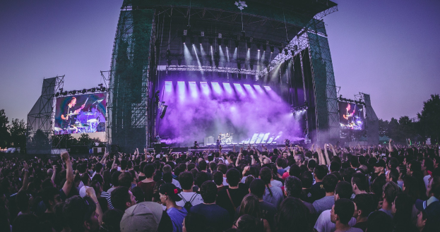 Eine große Menge bei einem Musikfestival schaut auf der Bühne mit Instrumenten und Bildschirmen, die vor einem Gebäude, Bäumen und einem blauen Himmel beleuchtet sind.