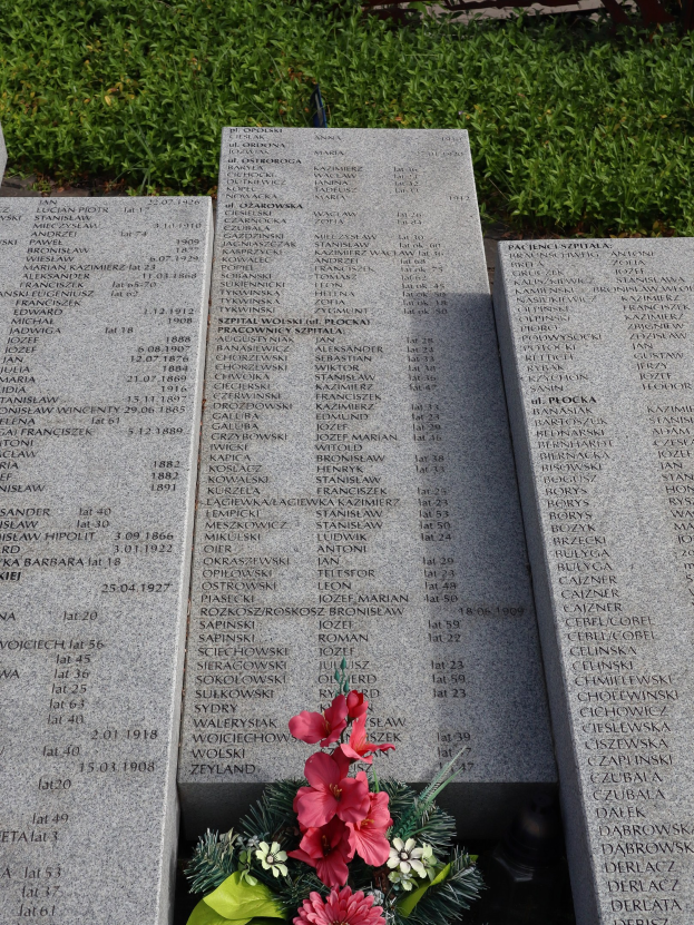 Gedenkstein mit einem Blumenstrauß darauf, umgeben von grünem Gras, mit Text, der wahrscheinlich Kriegsopfer gedenkt.