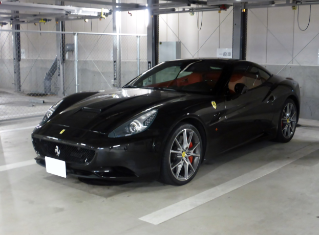 Schwarzer Ferrari California in einer Parkgarage mit einer Wand im Hintergrund, Maschendrahtzaun auf der linken Seite und Deckenbeleuchtung.