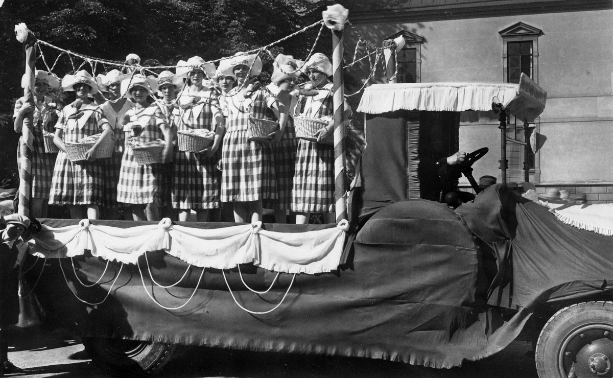 Schwarzes und weißes Foto einer Gruppe von Frauen, die auf einem Lkw stehen und Körbe halten, mit einem Gebäude und Bäumen im Hintergrund.