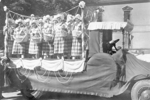 Schwarzes und weißes Foto einer Gruppe von Frauen, die auf einem Lkw stehen und Körbe halten, mit einem Gebäude und Bäumen im Hintergrund.