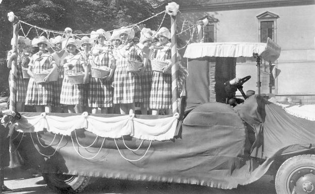 Schwarzes und weißes Foto einer Gruppe von Frauen, die auf einem Lkw stehen und Körbe halten, mit einem Gebäude und Bäumen im Hintergrund.
