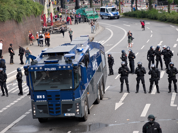 Gruppe von Polizisten vor einem gepanzerten Fahrzeug auf einer Stra├če, umgeben von bewaffneten Individuen mit Helmen, mit Geb├Ąuden, Verkehrszeichen und Pflanzen im Hintergrund.