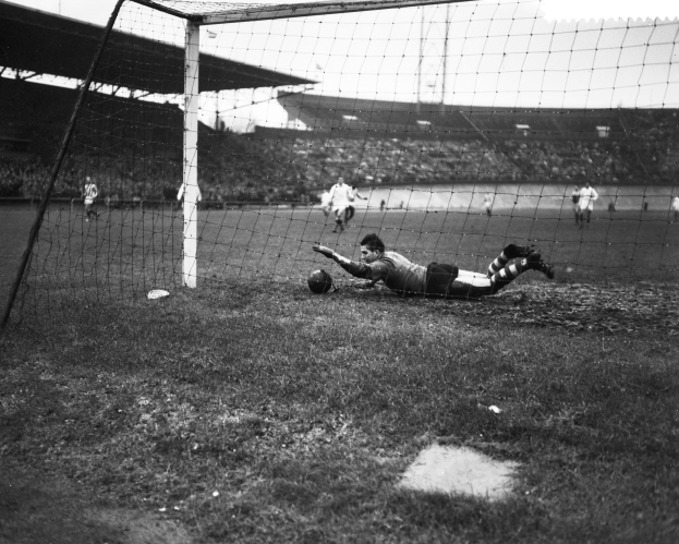 Ein Schwarz-Weiß-Foto eines Fussballspielers, der einen Ball vor einem Tor fangen will, umgeben von anderen Spielern auf dem Boden und einem Stadion mit Dach und Masten im Hintergrund bei bewölktem Himmel.