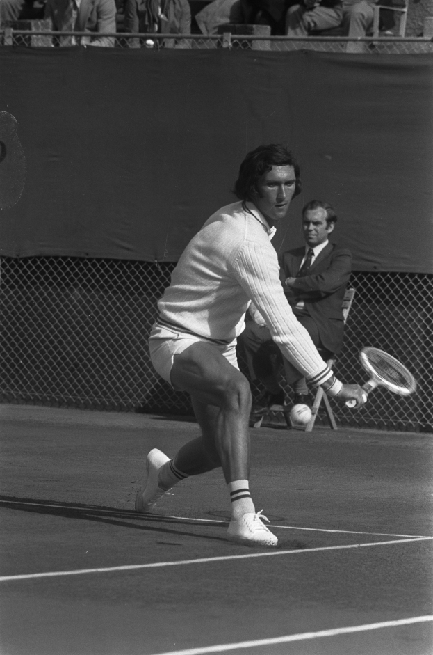 Ein professioneller Tennisspieler in Schuhen schwingt einen Schläger gegen einen Ball, mit Maschendrahtzaun und Zuschauern im Hintergrund.