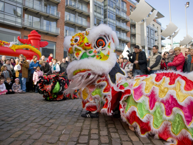 Ein lebendiges chinesisches Neujahrsfest in Amsterdam mit einem Löwen tanzen im Vordergrund mit einer Menge von Zuschauern, einige halten Kameras, vor einem Hintergrund von Gebäuden, Laternenmasten und einem klaren blauen Himmel.