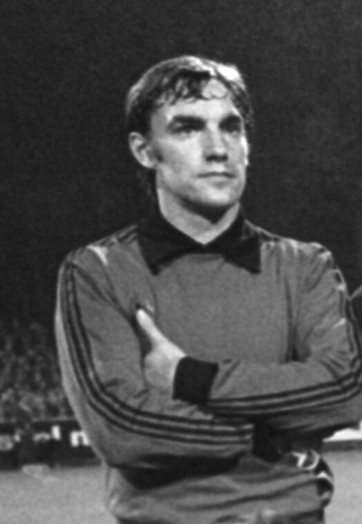 Schwarzes und weißes Foto eines Mannes in einem Anzug, der mit verschränkten Armen auf einem Fußballfeld steht und von einer Gruppe sitzender Menschen im Hintergrund umgeben ist.