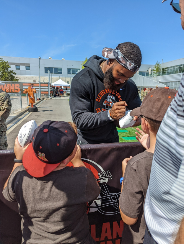 Cleveland Browns Wide Receiver Odell Beckham Jr. signiert Autogramme für Fans auf dem Trainingsgelände des Teams, umgeben von Menschen mit Kappen, einem Banner, Gebäuden, Bäumen und Zelten im Hintergrund bei einem bewölkten Himmel.