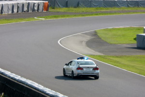 Ein Polizeiauto fährt auf einer Rennstrecke, umgeben von Gras mit Menschen, Zelten und anderen Gegenständen im Hintergrund.