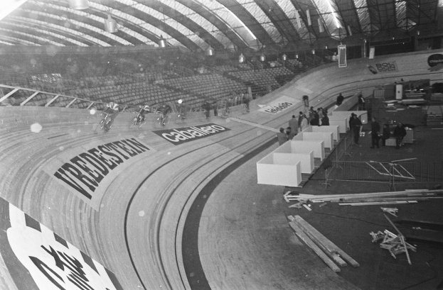 Ein Schwarz-Weiß-Foto eines Indoorstadions mit einer zentralen Laufbahn, einigen Menschen rechts, Stühlen links und einem beleuchteten Schuppen oben.