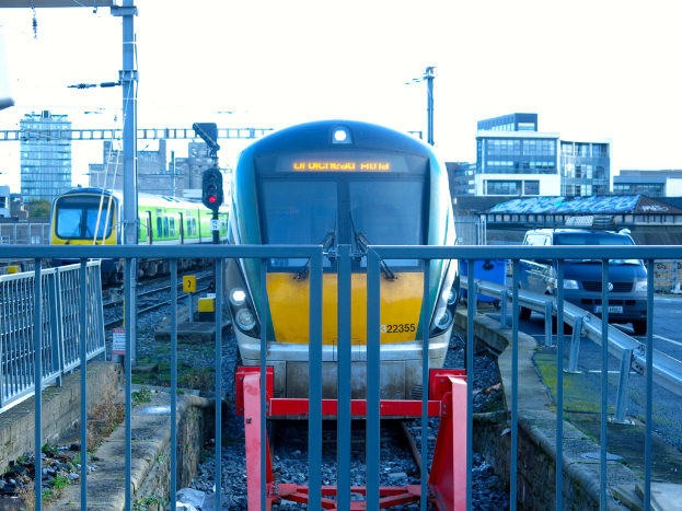 Zwei Züge auf dem Gleis mit einem Zaun im Vordergrund, Pfosten auf dem Land, einer mit einer Signalbeleuchtung, ein Auto auf der Straße rechts, Gebäude im Hintergrund und der Himmel darüber.