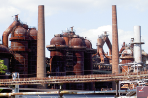 Ein großes Fabrikgebäude mit zahlreichen Rohren und Schornsteinen, einer Brücke, Masten, Lampen, Bäumen und einem bewölkten Himmel im Hintergrund.