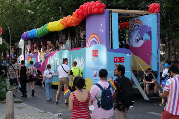 Eine Gruppe von Menschen, die auf einer Straße neben einem Lastwagen mit bunten Luftballons gehen, mit Schildern an der Straße und Bäumen und Gebäuden im Hintergrund, was auf eine Pride-Parade in Paris hindeutet.
