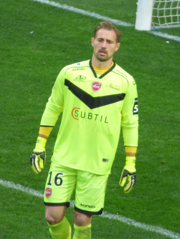 Ein Mann in einem gelben Torwarttrikot steht auf einem Fußballfeld mit einem Torpfosten im Hintergrund, trägt ein T-Shirt, Shorts, Socken und Handschuhe.