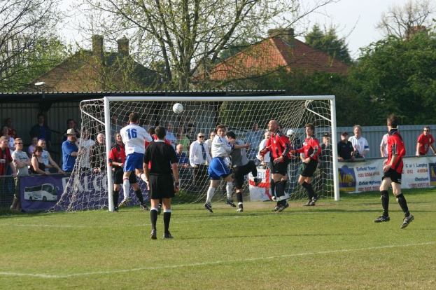Fussballspieler spielen auf einem Feld mit einem Tor, während Zuschauer dahinter stehen; im Hintergrund sind Bäume und Häuser zu sehen.
