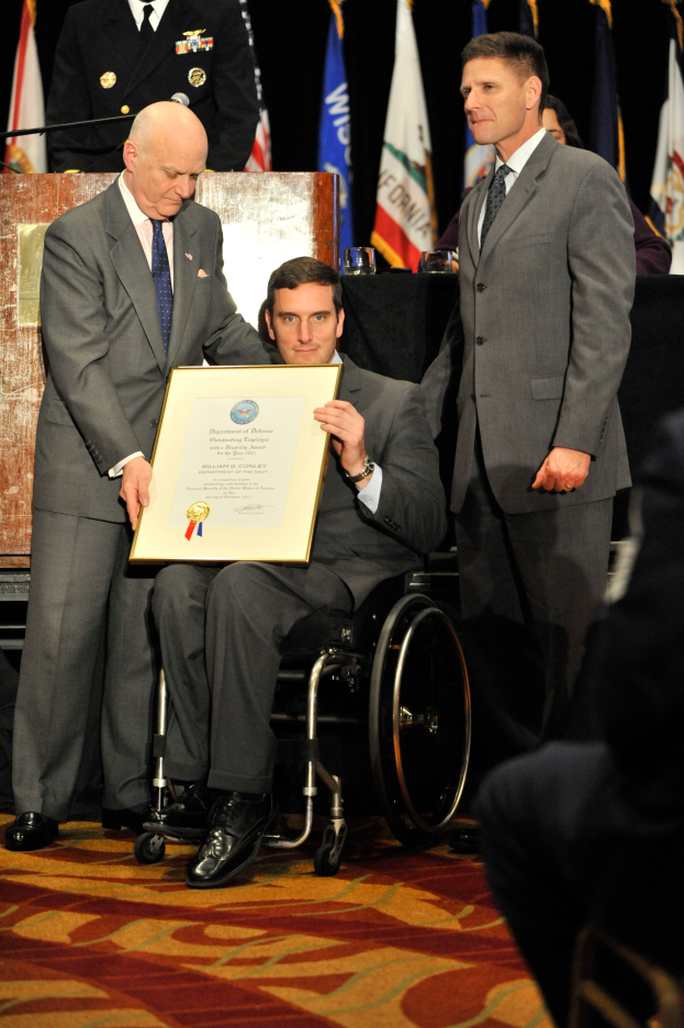Ein Mann im Rollstuhl erhält eine Auszeichnung von zwei Männern in Anzügen, die ein Zertifikat halten, mit Menschen, Flaggen und einem Podium im Hintergrund.