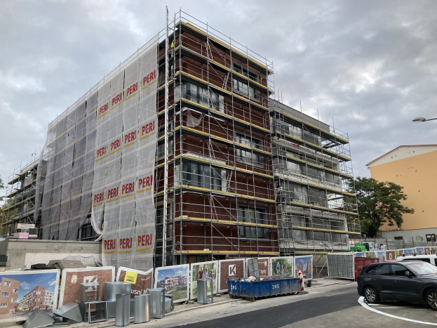 Großes Gebäude im Bau mit Gerüst, umgeben von Bäumen, Straßeninfrastruktur, Fahrzeugen und Werbetafeln unter einem bewölkten Himmel.