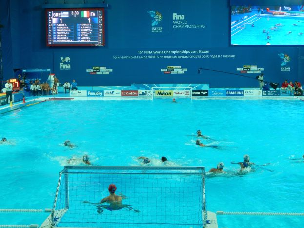 Gruppe von Athleten beim Wasserballspielen in einem Pool während der 2016 FINA-Weltmeisterschaften in Kazan, mit Zuschauern auf der linken Seite und einem Bildschirm mit Event-Branding im Hintergrund.