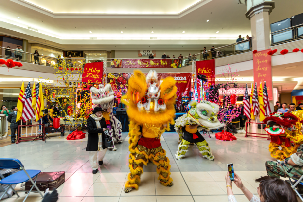 Ein lebendiger chinesischer Neujahrsfeier in einem Einkaufszentrum mit tanzenden Menschen in Kostümen und anderen mit Mobiltelefonen, umgeben von Fahnen, Bannern, Dekorationen und Lichtern.