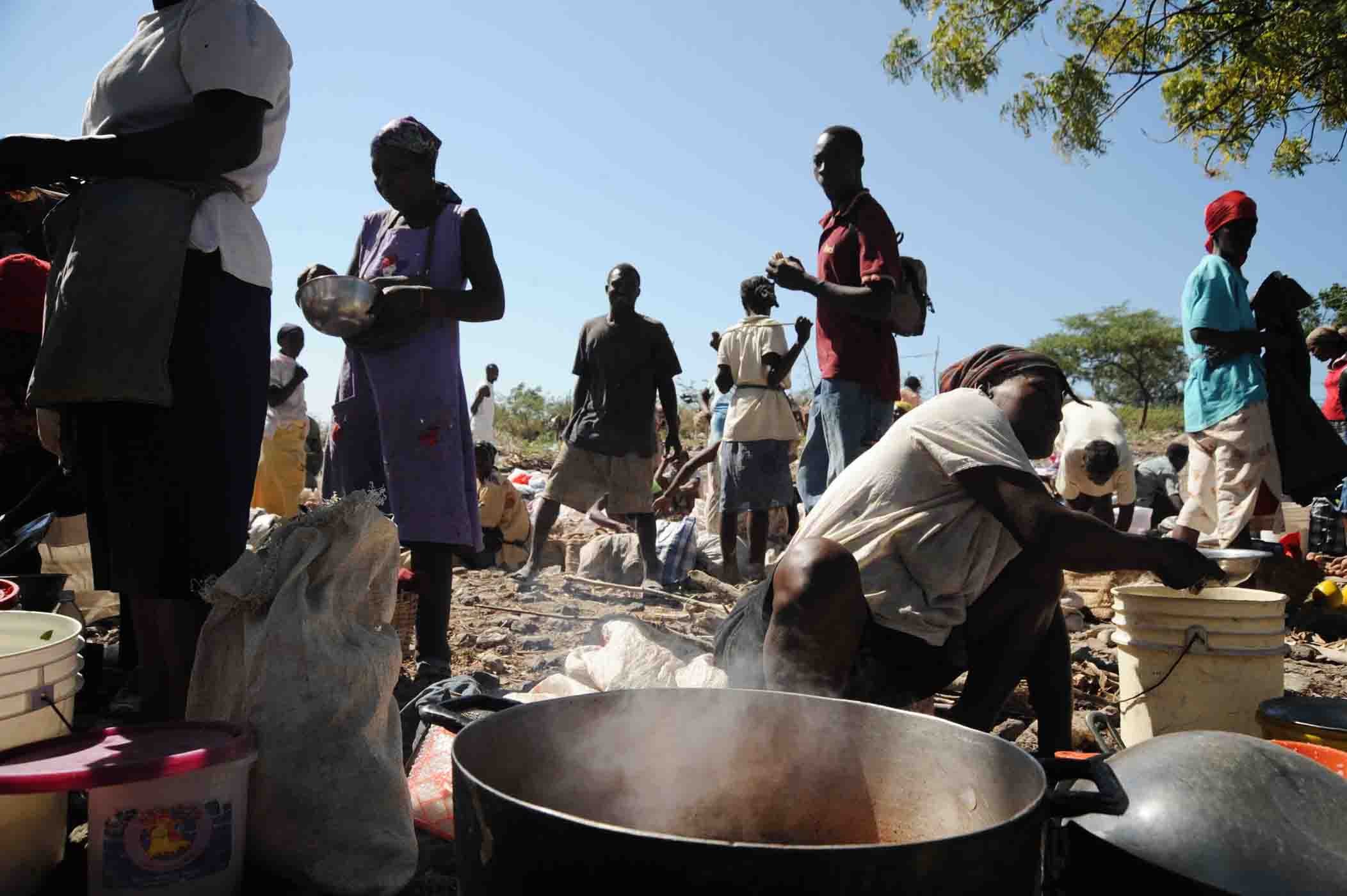 Menschen bereiten Essen im Freien in einer ländlichen Gegend zu, mit einem großen Topf umgeben von Behältern und Zubehör.