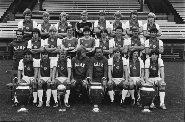 Ein Schwarz-Weiß-Foto einer grinsenden Fußballmannschaft, die mit ihren Trophäen posiert, einige sitzen auf Bänken und andere stehen, während die Trophäen vor ihnen auf dem Rasen platziert sind.