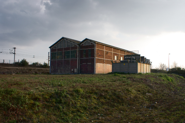 Eine Szene mit Schuppen, Transformatoren, Strommasten, Stromkabeln, Zäunen, einer Straßenlaterne an einem Laternenpfahl, Bäumen und einem bewölkten Himmel.